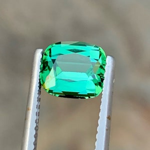 May include: A vibrant green gemstone, likely an emerald, is held in tweezers. The gemstone is rectangular in shape with a slight emerald cut.