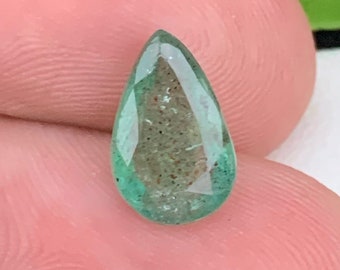 Puede incluir: Una piedra preciosa verde en forma de pera con inclusiones, probablemente una esmeralda, se sostiene en una mano.