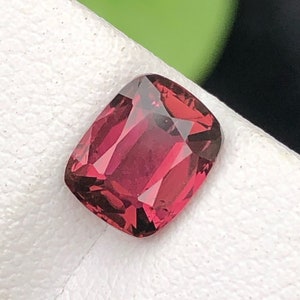 Pode incluir: Uma gema de cor vermelha profunda com corte almofada. A gema está colocada sobre um fundo branco.