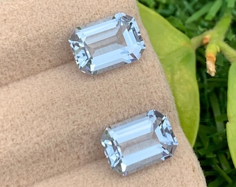 Op de afbeelding: Twee lichtblauwe smaragd geslepen edelstenen op een beige fluwelen kussen.