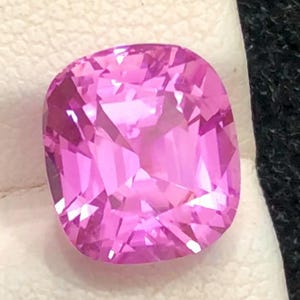 May include: A cushion-cut, faceted pink gemstone. The gem is a vibrant shade of pink, with a square shape and rounded corners. It is set against a white surface with a dark, textured background.