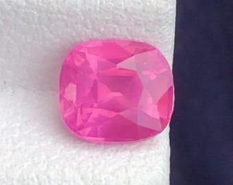 May include: A vibrant, cushion-cut, pink gemstone. The gem is set against a white textured surface, with a dark blue background. The gemstone's facets reflect light, showcasing its brilliance. This gemstone is likely for jewelry.