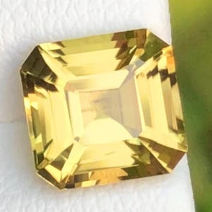 Puede incluir: Una gema amarilla tallada en forma cuadrada. La gema tiene forma rectangular con bordes biselados y una faceta cuadrada central. La gema está sobre un fondo blanco con un fondo borroso verde y amarillo.