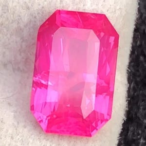 May include: A vibrant, rectangular-cut, hot pink gemstone. The gem is faceted, reflecting light and showcasing its clarity. The stone is set against a white and black background, highlighting its intense color and shape. This gemstone could be used in jewelry.