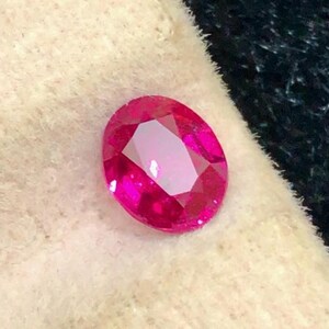 Può includere: Una gemma di rubino ovale e sfaccettata. La gemma è di un rosso vibrante con una superficie lucida, che riflette la luce. Il rubino è posto su uno sfondo chiaro e strutturato, con un tessuto scuro e strutturato sullo sfondo.