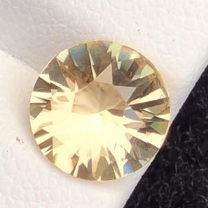 Puede incluir: Una gema redonda y facetada con un tono amarillo pálido. La gema está colocada sobre una superficie blanca y un fondo negro. Las facetas crean un efecto de estallido de estrella, reflejando la luz.