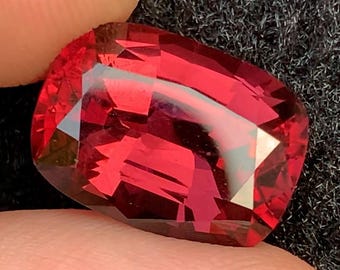May include: A faceted, oval-shaped, deep red gemstone. The gem is held between two fingers against a dark, textured background. The gem's surface reflects light, showcasing its cut and color.