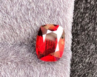 Puede incluir: Una gema roja facetada, de talla cojín, con una superficie brillante y reflectante. La gema se encuentra sobre un fondo gris suave. El color de la gema varía del rojo intenso a tonos más claros, mostrando su claridad y talla.
