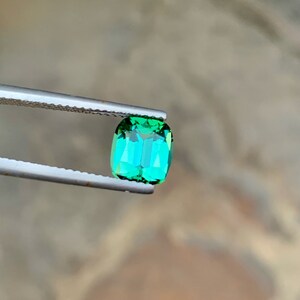 May include: A vibrant green gemstone with a rectangular cut, held by tweezers against a blurred background.