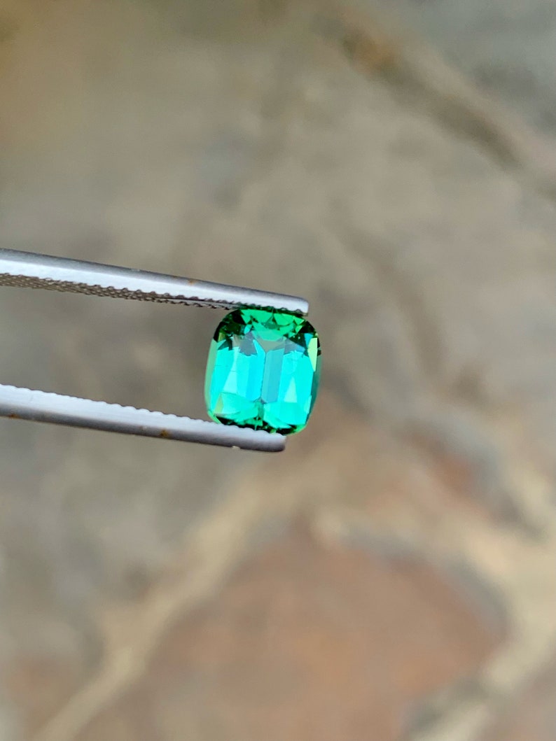 May include: A vibrant green gemstone with a rectangular cut, held by tweezers against a blurred background.