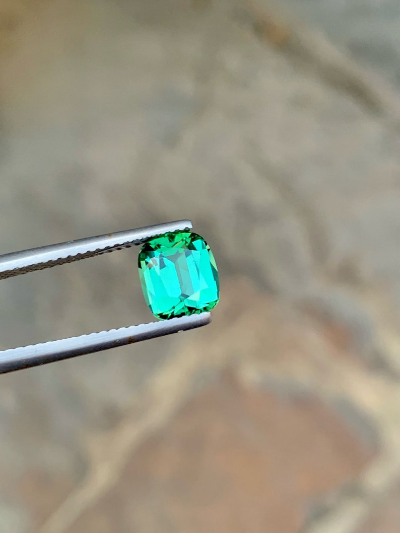 May include: A vibrant green gemstone, likely an emerald, is held in tweezers. The stone is oval-shaped and has a smooth, polished surface.