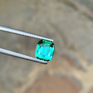May include: A vibrant green gemstone with a rectangular cut, held by tweezers against a blurred background.