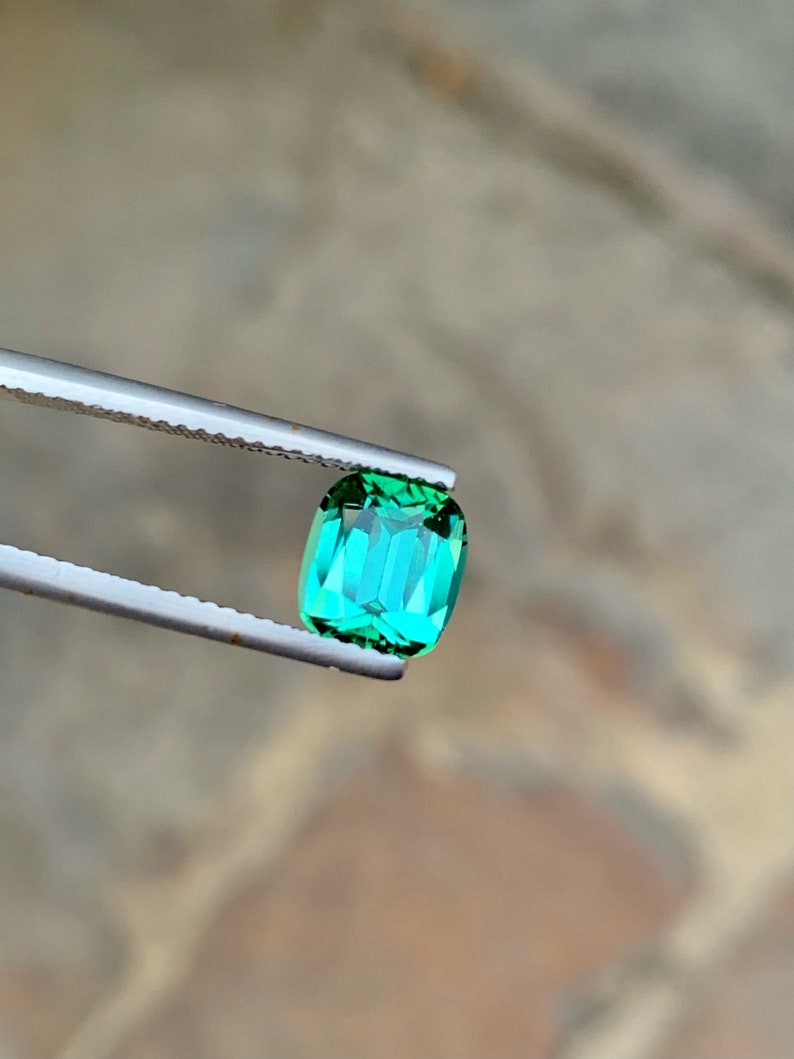 May include: A vibrant green gemstone with a rectangular cut, held by tweezers against a blurred background.