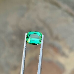 May include: A vibrant green gemstone, likely an emerald, is held in tweezers. The gemstone is rectangular in shape with a slight emerald cut.
