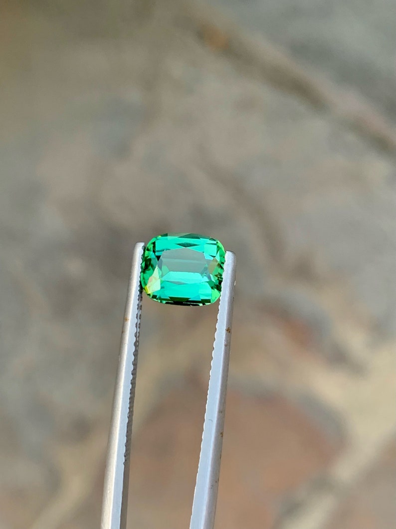 May include: A vibrant green gemstone, likely an emerald, is held in tweezers. The gemstone is rectangular in shape with a slight emerald cut.