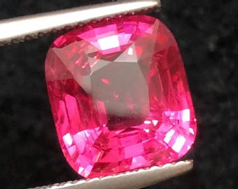 May include: A vibrant, cushion-cut, hot pink gemstone is held by a pair of tweezers against a dark background. The gemstone's facets reflect light, creating a sparkling effect. This precious stone is likely for jewelry.