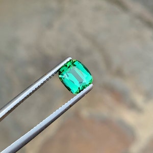 May include: A vibrant green gemstone with a rectangular cut, held by tweezers against a blurred background.