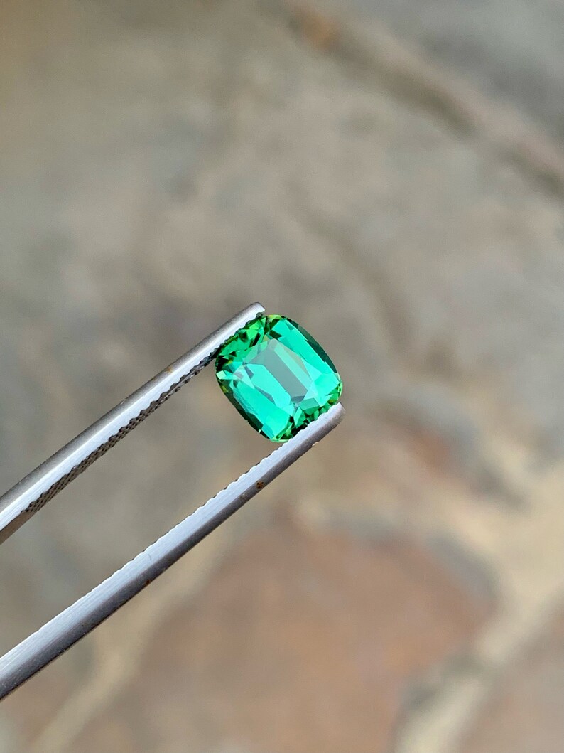 May include: A vibrant green gemstone with a rectangular cut, held by tweezers against a blurred background.