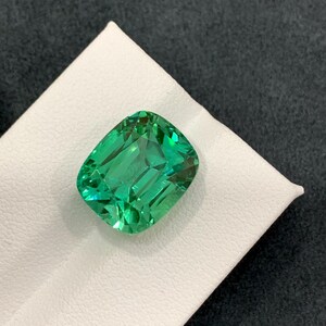 May include: A vibrant, emerald-green gemstone with a rectangular cushion cut. The gem is displayed on a white surface against a dark gray background, showcasing its facets and brilliance. This gemstone is a beautiful example of fine jewelry.