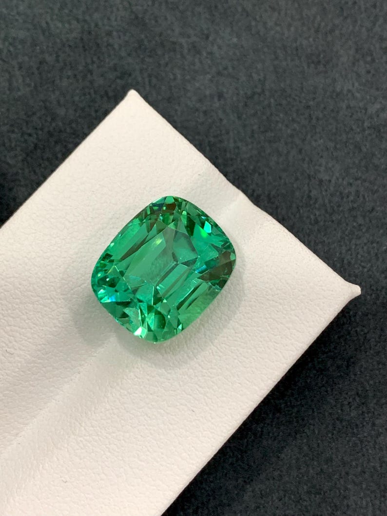 May include: A vibrant, emerald-green gemstone with a rectangular cushion cut. The gem is displayed on a white surface against a dark gray background, showcasing its facets and brilliance. This gemstone is a beautiful example of fine jewelry.