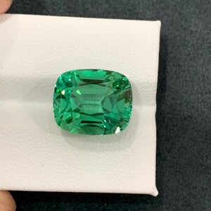 May include: A vibrant, emerald-cut green gemstone. The gem is set on a white surface, showcasing its clarity and brilliance. The stone's facets reflect light, creating a sparkling effect. The gemstone is a striking example of fine jewelry.
