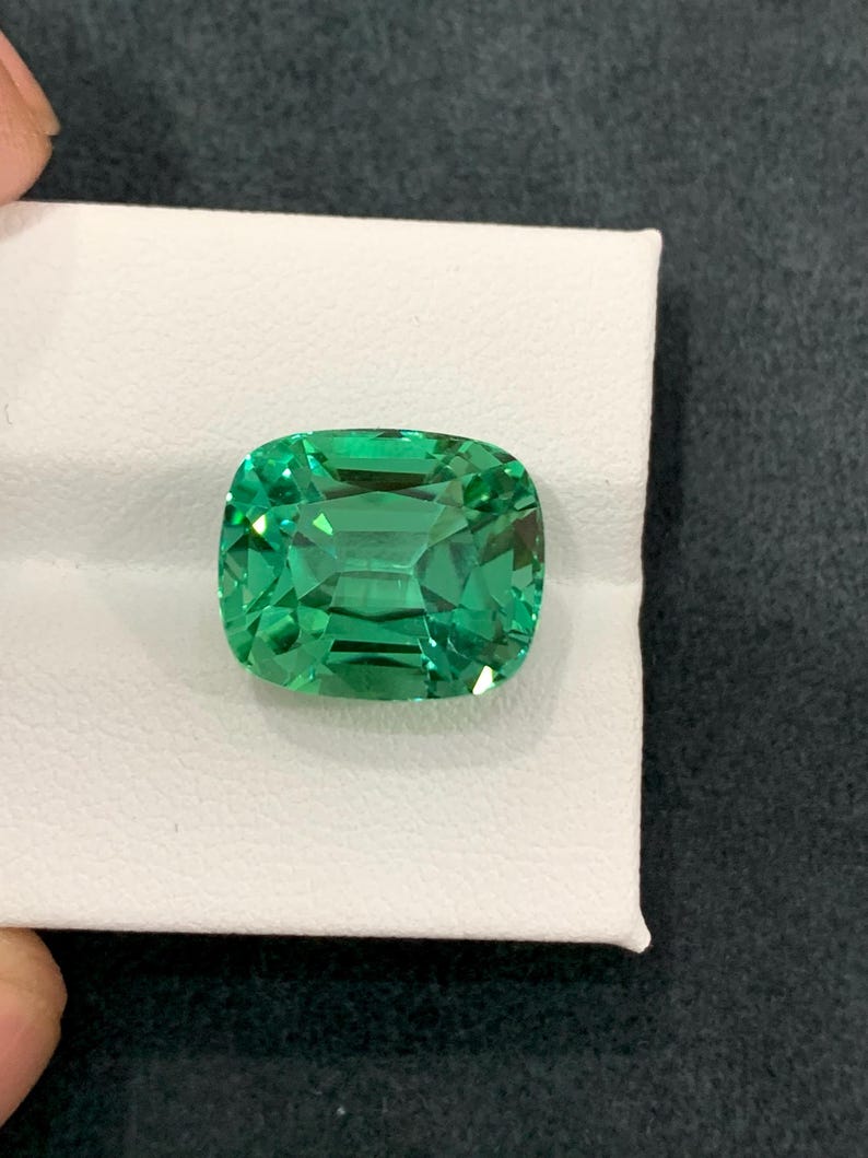 May include: A vibrant, emerald-cut green gemstone. The gem is set on a white surface, showcasing its clarity and brilliance. The stone's facets reflect light, creating a sparkling effect. The gemstone is a striking example of fine jewelry.