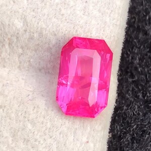 May include: A vibrant, rectangular-cut, hot pink gemstone. The gem has a faceted surface that reflects light, creating a brilliant display. The stone is set against a white and black background.