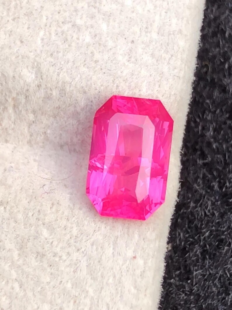 May include: A vibrant, rectangular-cut, hot pink gemstone. The gem has a faceted surface that reflects light, creating a brilliant display. The stone is set against a white and black background.