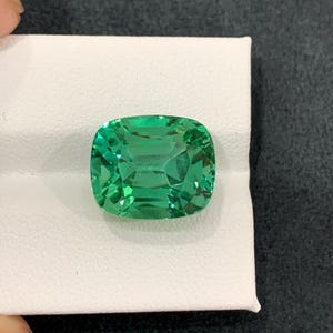 May include: A vibrant, emerald-green gemstone with a cushion cut. The gem is set on a white surface, showcasing its clarity and brilliance. The stone's facets reflect light, creating a captivating display of color and sparkle. A beautiful piece for jewelry.