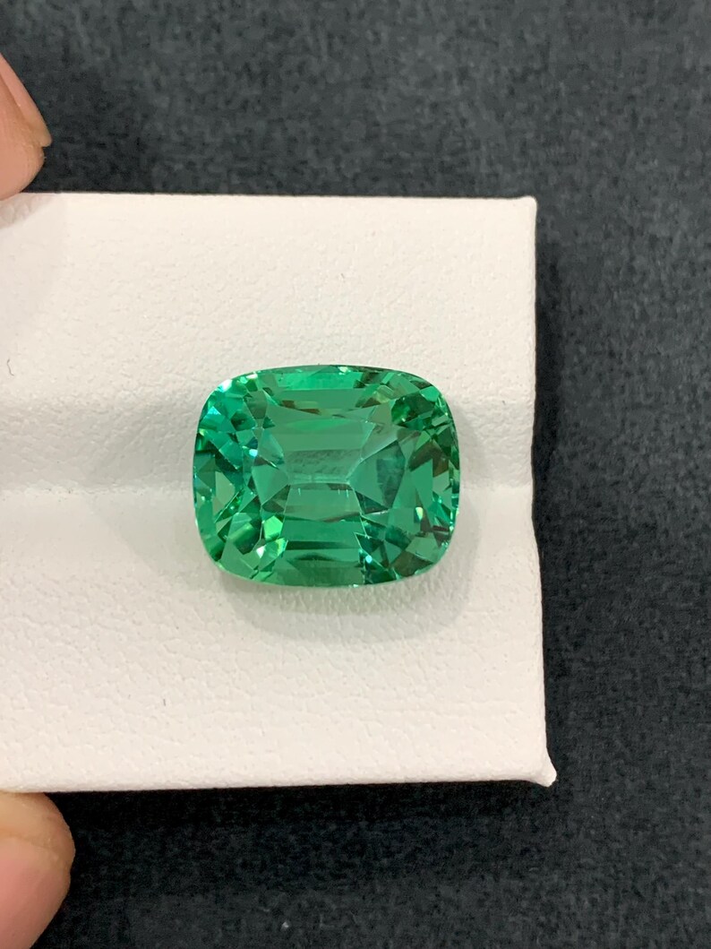 May include: A vibrant, emerald-green gemstone with a cushion cut. The gem is set on a white surface, showcasing its clarity and brilliance. The stone's facets reflect light, creating a captivating display of color and sparkle. A beautiful piece for jewelry.