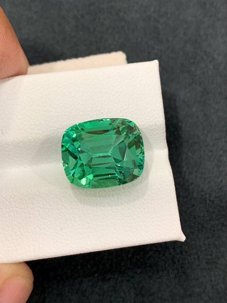 May include: A vibrant, emerald-cut gemstone with a brilliant green hue. The gem is held on a white surface, showcasing its facets and clarity. The stone's rectangular shape with rounded corners is prominent.
