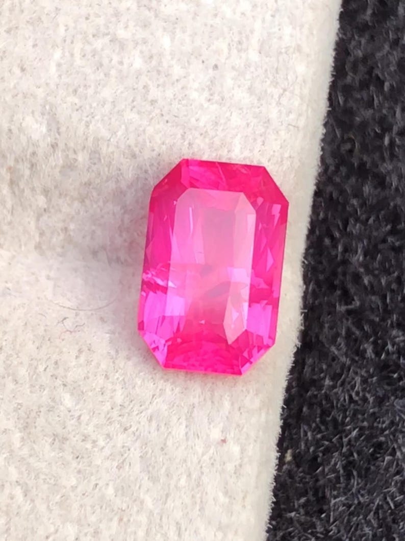 May include: A vibrant, rectangular-cut, hot pink gemstone. The gem is faceted, reflecting light and showcasing its clarity. The stone is set against a white and black background, highlighting its intense color and shape. This gemstone could be used in jewelry.