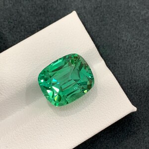 May include: A vibrant, emerald-green gemstone with a cushion cut. The gem is displayed on a white surface against a dark background. The facets of the stone reflect light, creating a sparkling effect. This gemstone is likely for jewelry.
