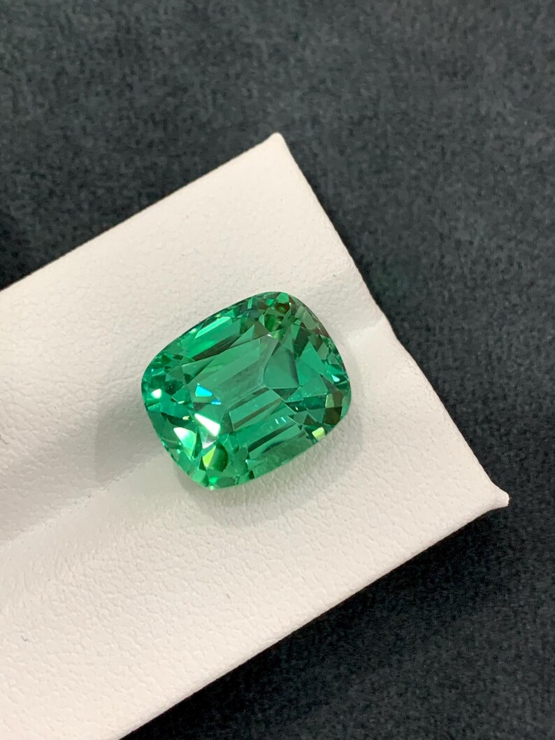 May include: A vibrant, emerald-green gemstone with a cushion cut. The gem is displayed on a white surface against a dark background. The facets of the stone reflect light, creating a sparkling effect. This gemstone is likely for jewelry.