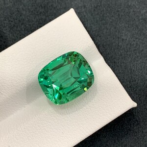 May include: A vibrant, emerald-green gemstone with a cushion cut. The gem is displayed on a white surface against a dark gray background. The facets of the stone reflect light, showcasing its clarity and brilliance. This gemstone is a beautiful example of fine jewelry.