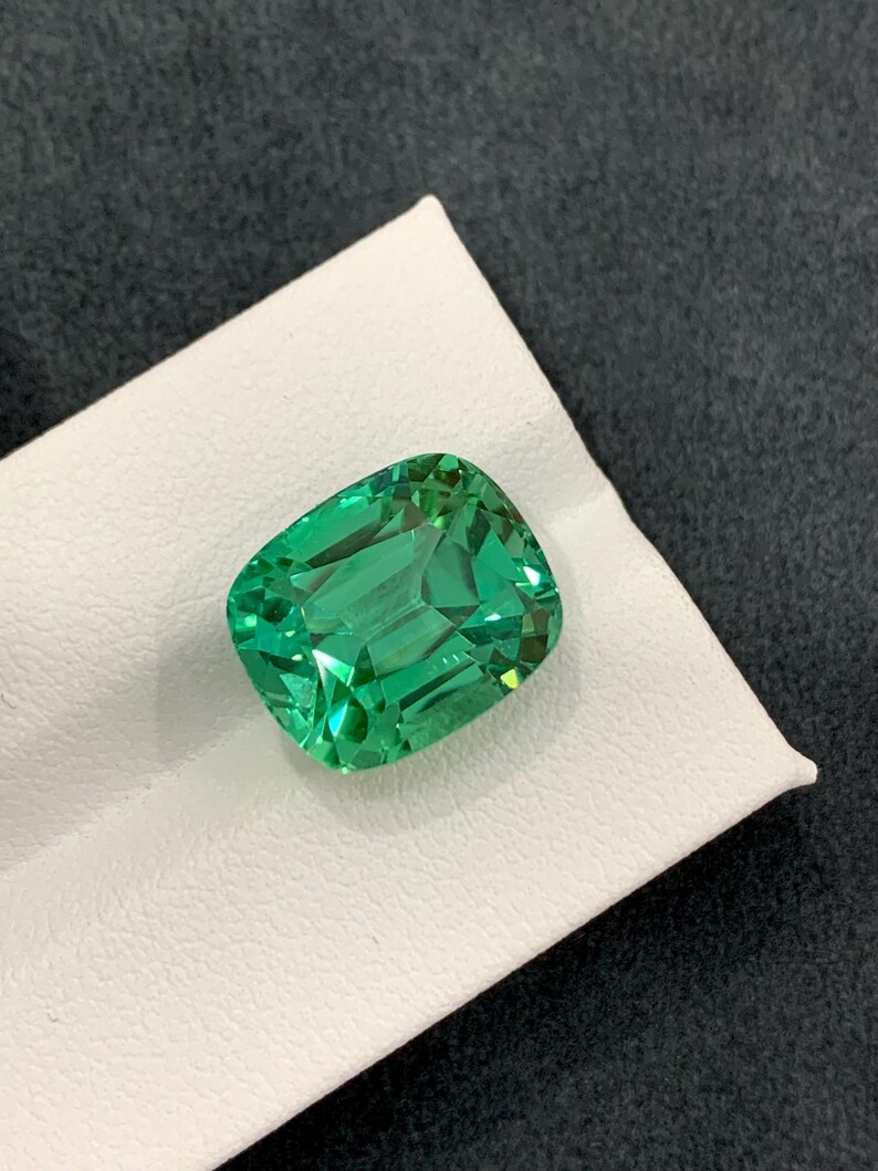 May include: A vibrant, emerald-green gemstone with a cushion cut. The gem is displayed on a white surface against a dark gray background. The facets of the stone reflect light, showcasing its clarity and brilliance. This gemstone is a beautiful example of fine jewelry.