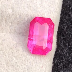 May include: A vibrant, rectangular-cut, hot pink gemstone. The gem is faceted, catching the light and displaying a bright, clear color. The gemstone is set against a white and black background.