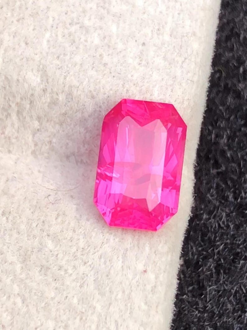 May include: A vibrant, rectangular-cut, hot pink gemstone. The gem is faceted, catching the light and displaying a bright, clear color. The gemstone is set against a white and black background.