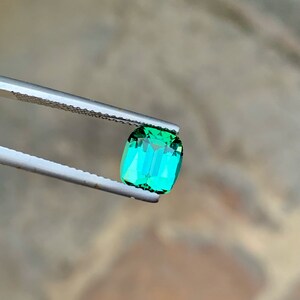 May include: A vibrant green gemstone with a rectangular cut, held by tweezers against a blurred background.