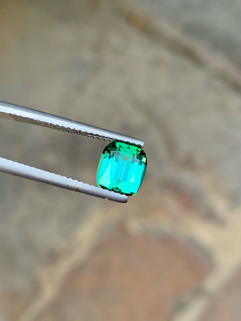 May include: A vibrant green gemstone with a rectangular cut, held by tweezers against a blurred background.