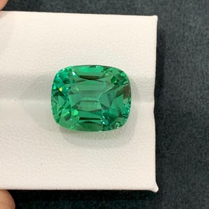 May include: A vibrant, cushion-cut gemstone with a bright green hue. The gem is set on a white surface, showcasing its facets and internal reflections. The stone's shape is square with rounded corners, reflecting light.