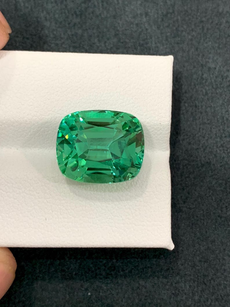 May include: A vibrant, cushion-cut gemstone with a bright green hue. The gem is set on a white surface, showcasing its facets and internal reflections. The stone's shape is square with rounded corners, reflecting light.