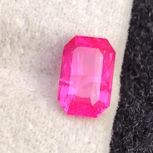 May include: A vibrant, rectangular-cut, hot pink gemstone. The gem is set against a white and black textured background. The gemstone's facets reflect light, creating a brilliant display of color.
