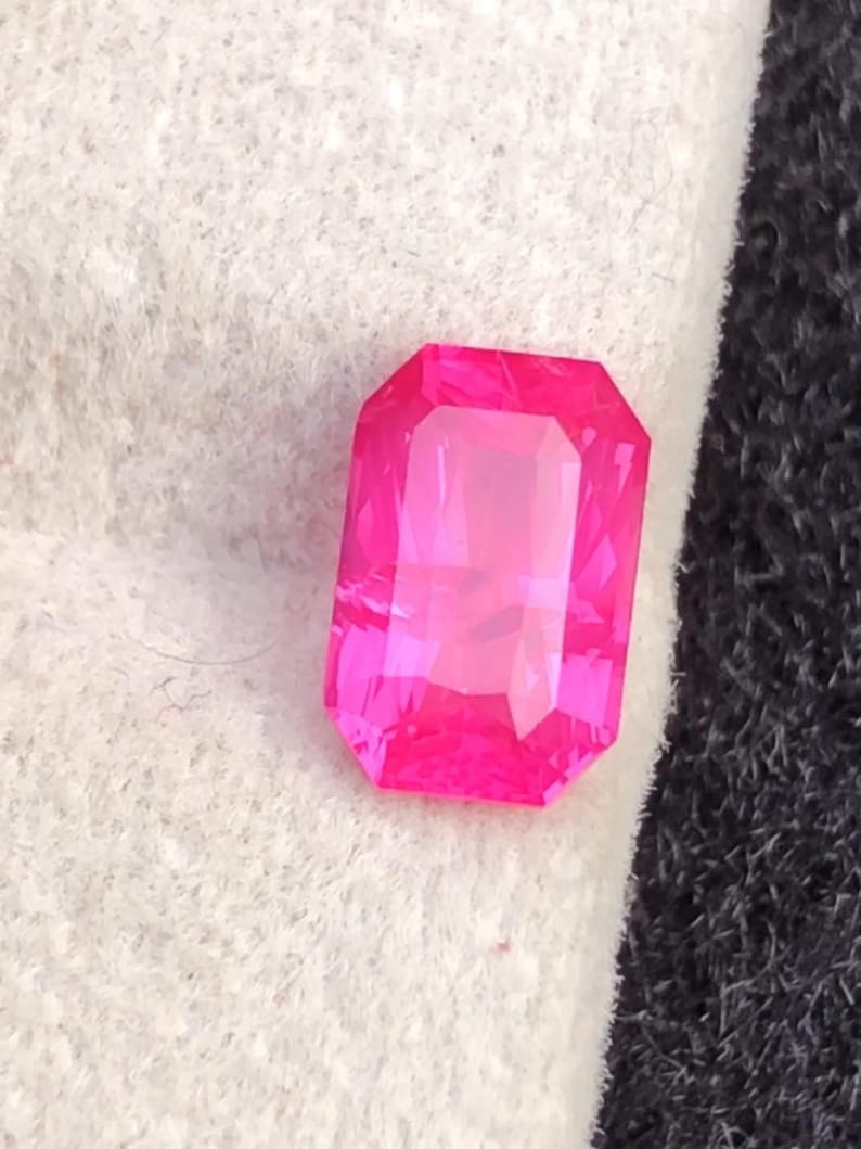 May include: A vibrant, rectangular-cut, hot pink gemstone. The gem is set against a white and black textured background. The gemstone's facets reflect light, creating a brilliant display of color.
