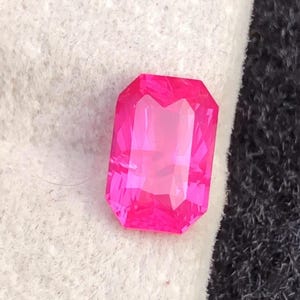 May include: A vibrant, rectangular-cut, hot pink gemstone with multiple facets. The gem is set against a white and black background, showcasing its brilliant color and clarity. The gemstone's shape is a classic emerald cut.