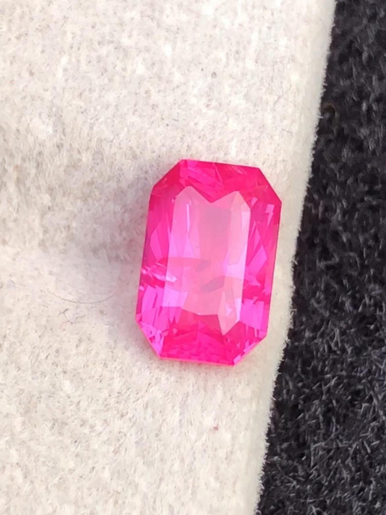 May include: A vibrant, rectangular-cut, hot pink gemstone with multiple facets. The gem is set against a white and black background, showcasing its brilliant color and clarity. The gemstone's shape is a classic emerald cut.