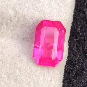 May include: A vibrant, rectangular-cut, hot pink gemstone. The gem has a faceted surface that reflects light, creating a brilliant shine. The stone is set against a white and black background.
