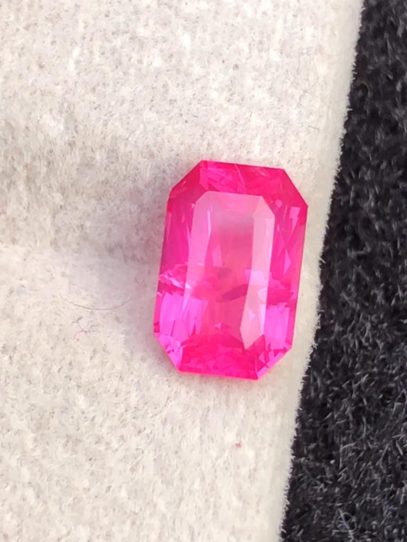 May include: A vibrant, rectangular-cut, hot pink gemstone. The gem has a faceted surface that reflects light, creating a brilliant shine. The stone is set against a white and black background.