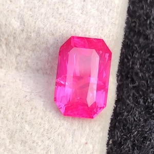 May include: A vibrant, rectangular-cut, hot pink gemstone. The gem has a faceted surface that reflects light, creating a brilliant shine. The stone is set against a white and black background, highlighting its color and shape. This gemstone is suitable for jewelry.