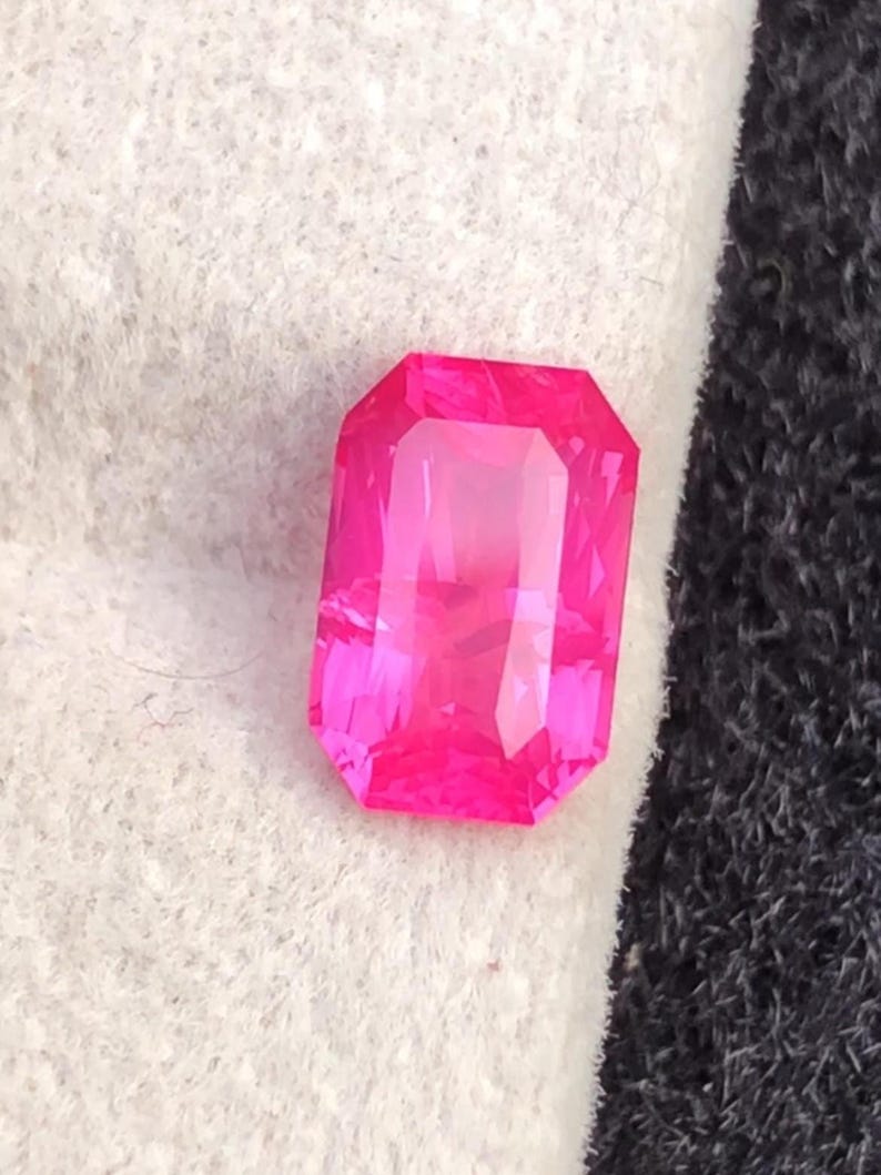 May include: A vibrant, rectangular-cut, hot pink gemstone. The gem has a faceted surface that reflects light, creating a brilliant shine. The stone is set against a white and black background, highlighting its color and shape. This gemstone is suitable for jewelry.